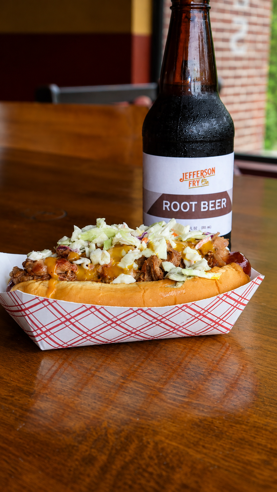 A loaded hot dog paired with a Jefferson Fry Co. root beer bottle on a wooden table.